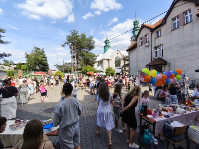 Uczniowie i społeczność lokalna na placu szkolnym podczas rodzinnego festynu. Naokoło placu stoiska ze smakołykami. W tle budynek kościoła oraz fasada szkoły.